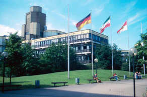 Altes Physikgebäude Altes Physikgebäude: Ein braun-gelbes Gebäude mit drei Stockwerken und einem hohen Turm links hinten. Davor eine Wiese und ein Platz mit Bänken und vier Fahnenmasten, an denen eine Deutschland-Fahne, eine NRW-Fahne und eine Dortmund-Fahne.