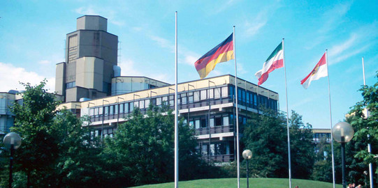 Altes Physikgebäude Altes Physikgebäude: Ein braun-gelbes Gebäude mit drei Stockwerken und einem hohen Turm links hinten. Davor eine Wiese und ein Platz mit Bänken und vier Fahnenmasten, an denen eine Deutschland-Fahne, eine NRW-Fahne und eine Dortmund-Fahne.